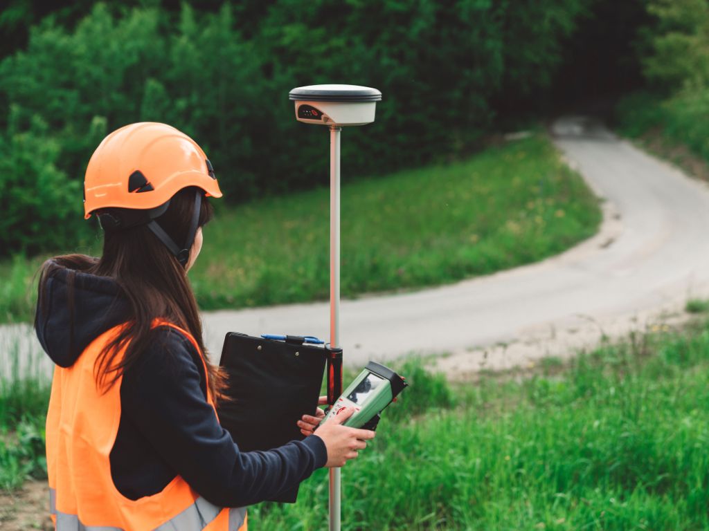 Frau mit GNSS-Rover und Tablet bei Vermessung auf Wiese neben Landstraße